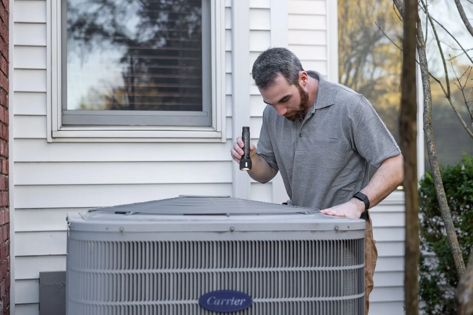 An HVAC technician inspects an outdoor air conditioning unit with a flashlight